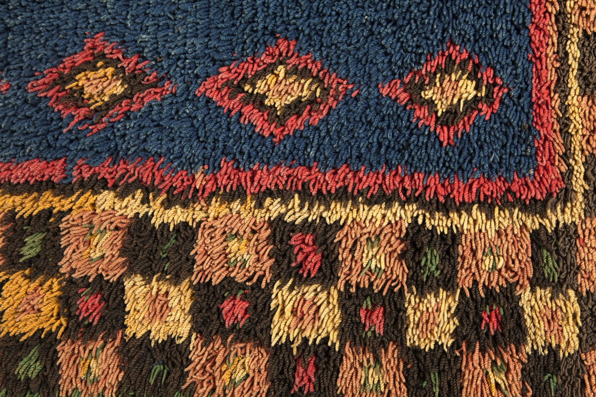 Prydnadsrya i två våder, hopsydda kant i kant. Botten i korndräll med varp av blått s-tvinnat lingarn 11 tr/cm och inslag av mörkbrun s-spunnen ull 7½ inslag/cm. Nockor av en tråd tvåtrådigt z-tvinnat hårdtvinnat ullgarn, knutna med giordesknut över 4 varptrådar med 4 varptrådar mellan nockorna och 4 inslag mellan nockraderna. Nockorna syns ej på baksidan.

Mönster: Stort blått mittfält avgränsat mot bården av en röd, en grönbrun och en gul smal linje. På det blå mittfältet, utmed sidorna romber av rött, grönbrunt och gult. Bred bård av schackrutor i grönbrunt, gult och rosagult. Gröna, röda och gula prickar i rutorna. Ytterst smal grön rand.