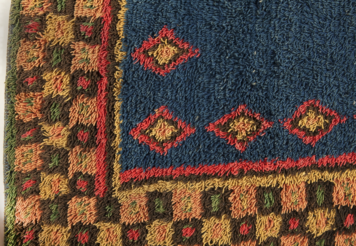 Prydnadsrya i två våder, hopsydda kant i kant. Botten i korndräll med varp av blått s-tvinnat lingarn 11 tr/cm och inslag av mörkbrun s-spunnen ull 7½ inslag/cm. Nockor av en tråd tvåtrådigt z-tvinnat hårdtvinnat ullgarn, knutna med giordesknut över 4 varptrådar med 4 varptrådar mellan nockorna och 4 inslag mellan nockraderna. Nockorna syns ej på baksidan.

Mönster: Stort blått mittfält avgränsat mot bården av en röd, en grönbrun och en gul smal linje. På det blå mittfältet, utmed sidorna romber av rött, grönbrunt och gult. Bred bård av schackrutor i grönbrunt, gult och rosagult. Gröna, röda och gula prickar i rutorna. Ytterst smal grön rand.