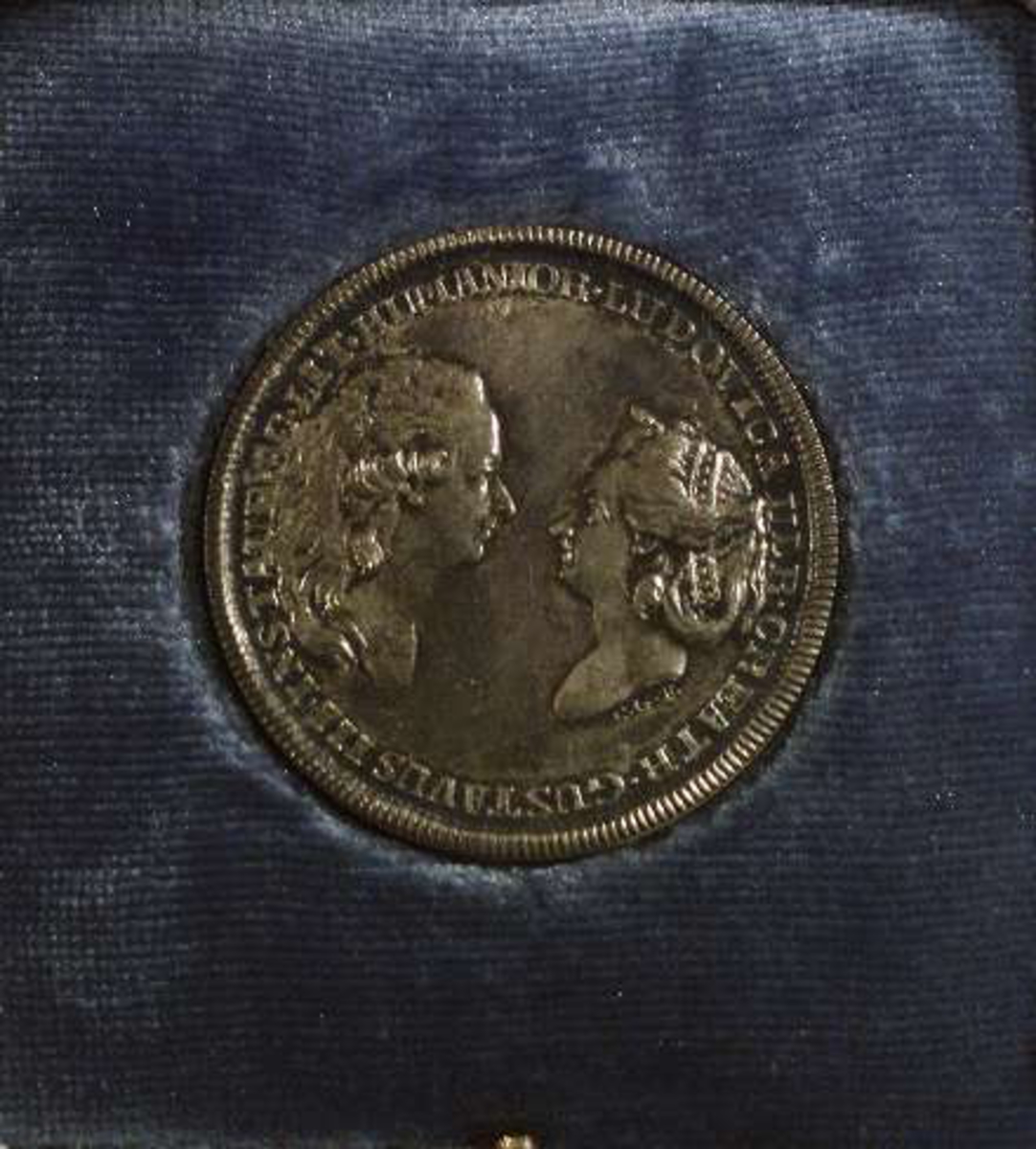Jettong av silver. På åtsidan i relief: Ett mans- och ett kvinnohuvud i profil vända mot varandra. Text: LUDOVICA ULR CREATR GUSTAVUS III INSTAUR AC LIT HUMANIOR (Gustaf III och Lovisa Ulrika). På jettongens frånsida i relief: Altare eller solur med ljusstrålar över. Text: LUCIS METITUR PROGRESSIBUS. På jettongens kantsida inristad liten text: SILVER (otydligt) 1929. 