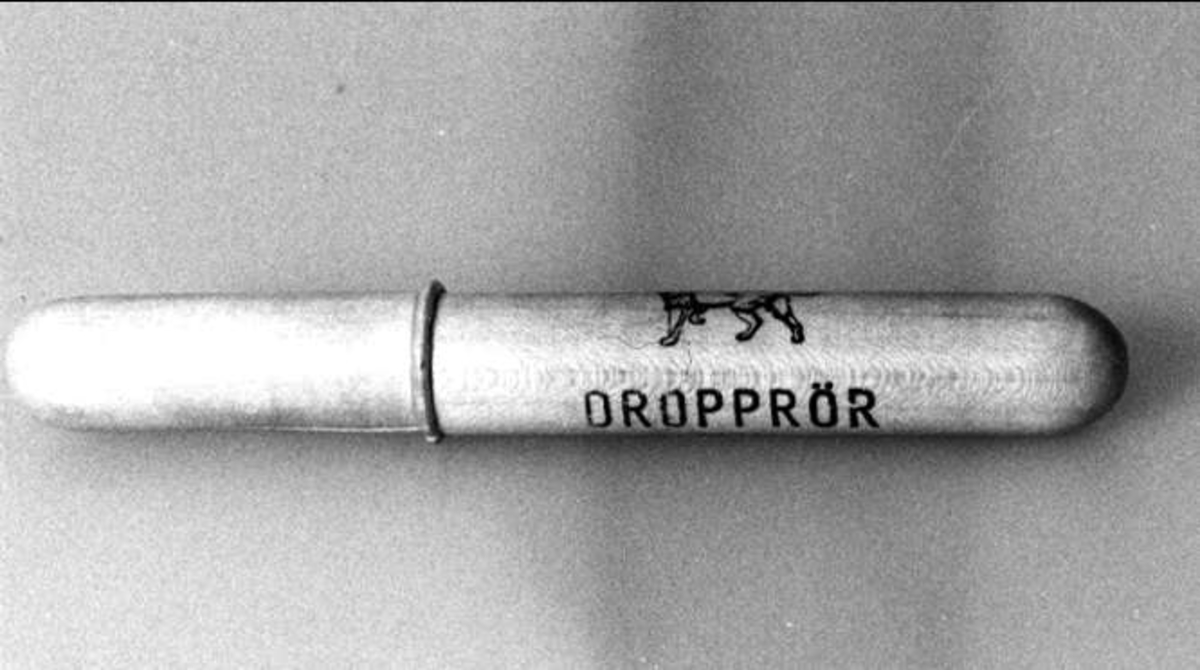 Förpackning för dropprör. Rörformigt i ljust lackerat trä. Två stycken som träs i varandra. Ett tryckt lejon i svart. Därunder i svart tryckt text: DROPPRÖR 7702 c, Apoteket Lejonet, Upsala. Stora Torget 5. 