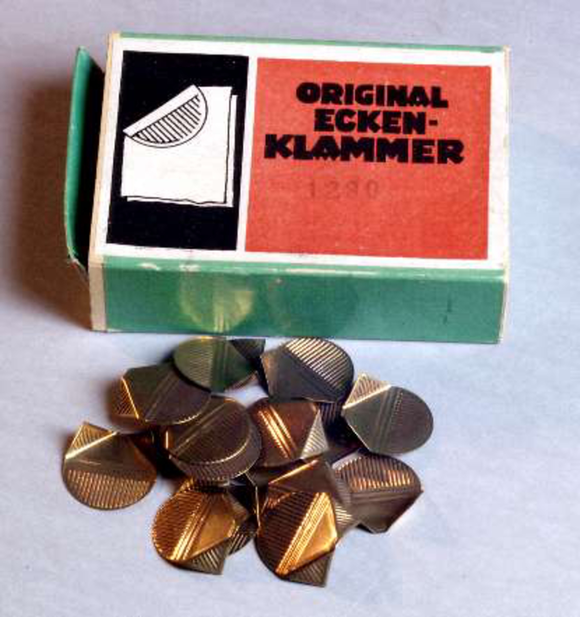 Skjutask av papp klädd med grönt papper. På ovansidan pappersetikett i rött och svart med text: ORIGINAL ECKEN-KLAMMER, 1280 och bild av hur en klämma viks runt hörnet på ett papper.
Innehåller mässingsbleck med vikmarkeringar och räfflor, se UM22320b.