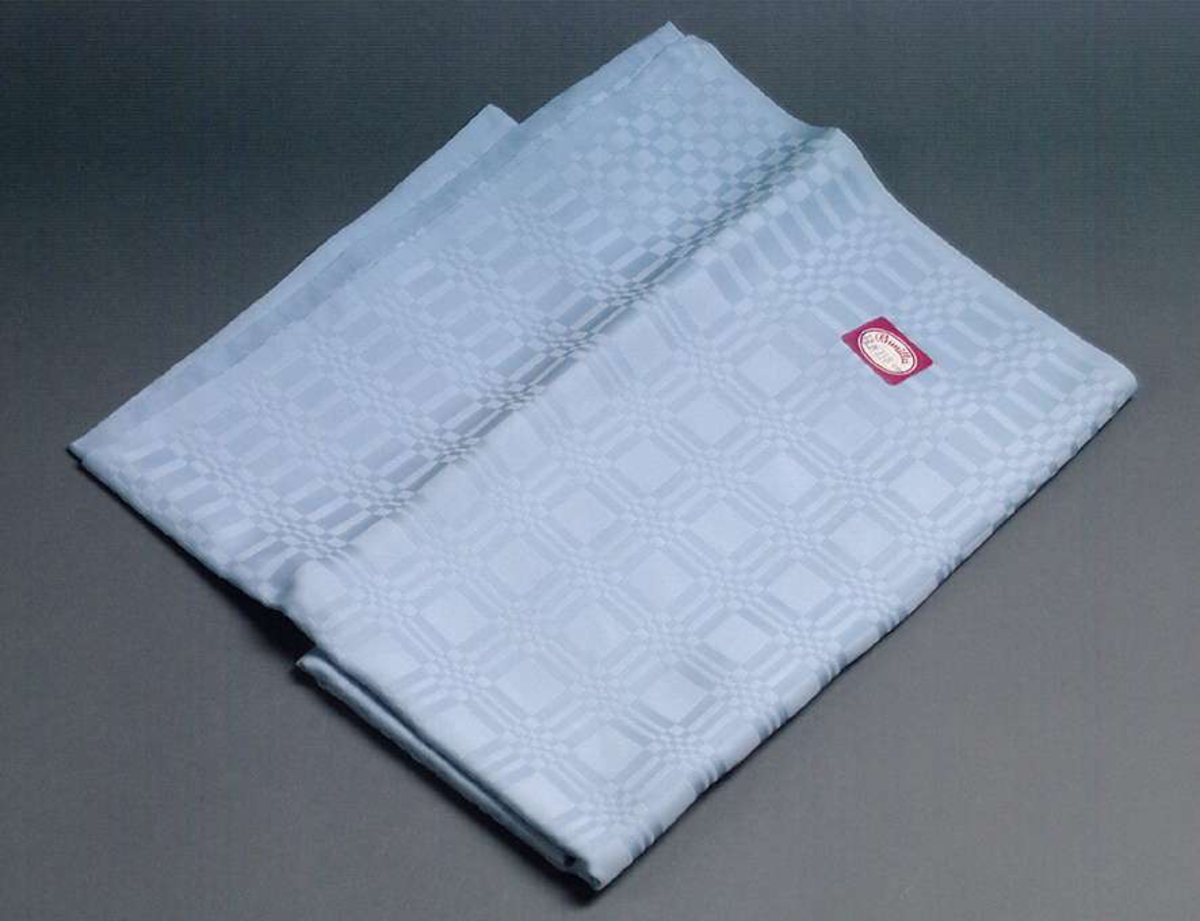 Ljusblå kvadratisk bordsduk av perlon i drällteknik med stadkanter runtom. Dukens mått: 115 x 115 cm. Påklistrad pappersetikett i rött och vitt med texten: Gunilla samt måtten.