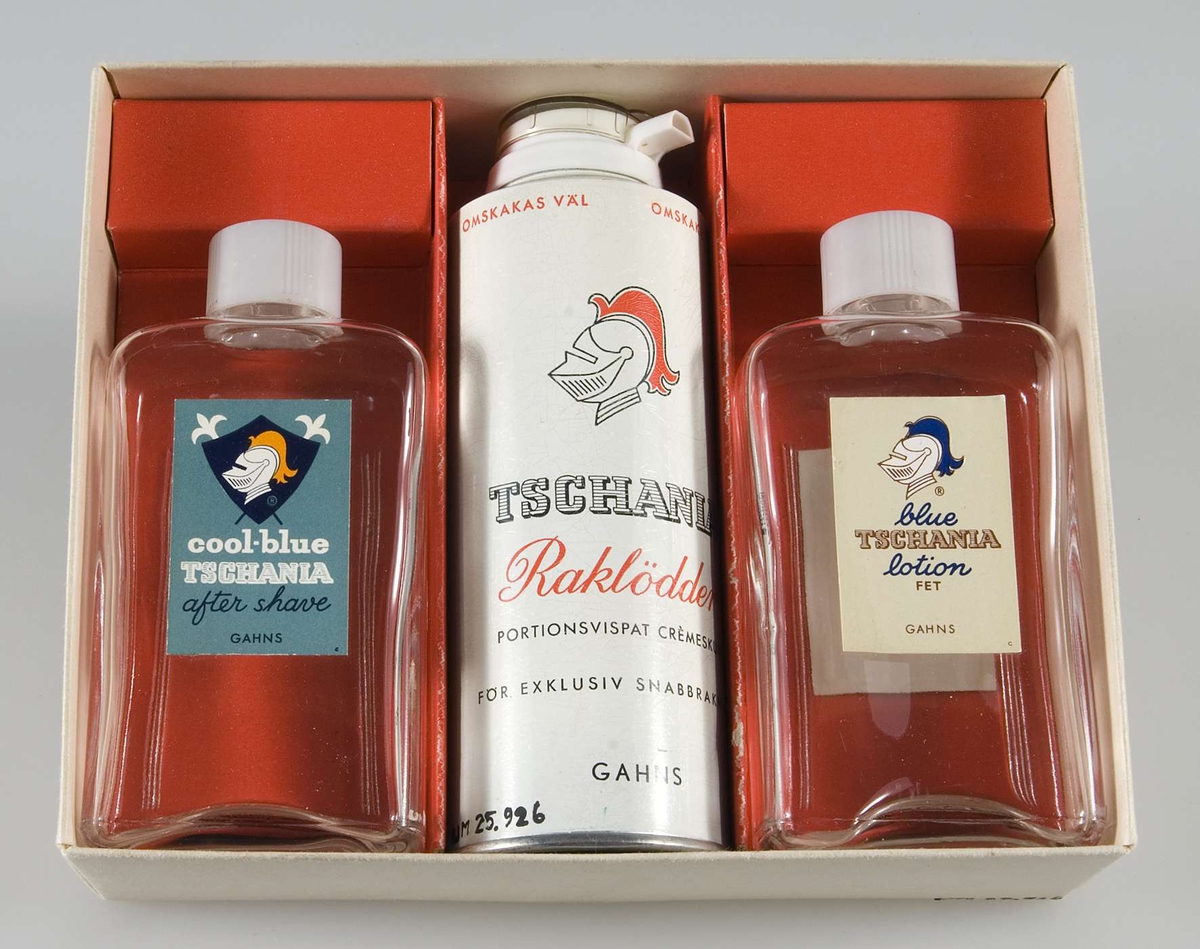 Raklödder och rakvatten i presentförpackning. Pappask i gulvitt med guldband och svart och vit text : TSCHANIA, För exklusiv atmosfär, GAHNS och bild av herrar i frack och hög hatt. Innehållande två rektangulära glasflaskor av ofärgat glas med vitt plastlock och pappersetikett med text: Cool-Blue Tschania after shave Gahns, resp: Blue Tschania Lotion, vilande på röd papp samt i mitten en sprayflaska av vitlackerad plåt med text: Tshania Raklödder Gahns.