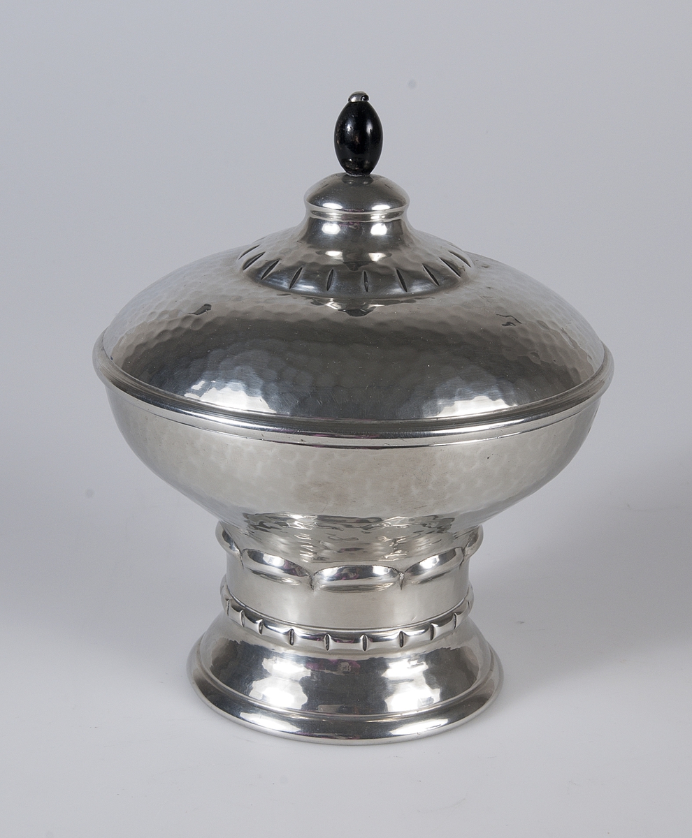 Skål med lock av hamrat tenn. Locket har en svart knopp, eventuellt av trä. Skålen har dekorerat fotparti. Stämplad: MAGS TIN. Ingraverat: Intresserad Thorares Hederspris 1927.

