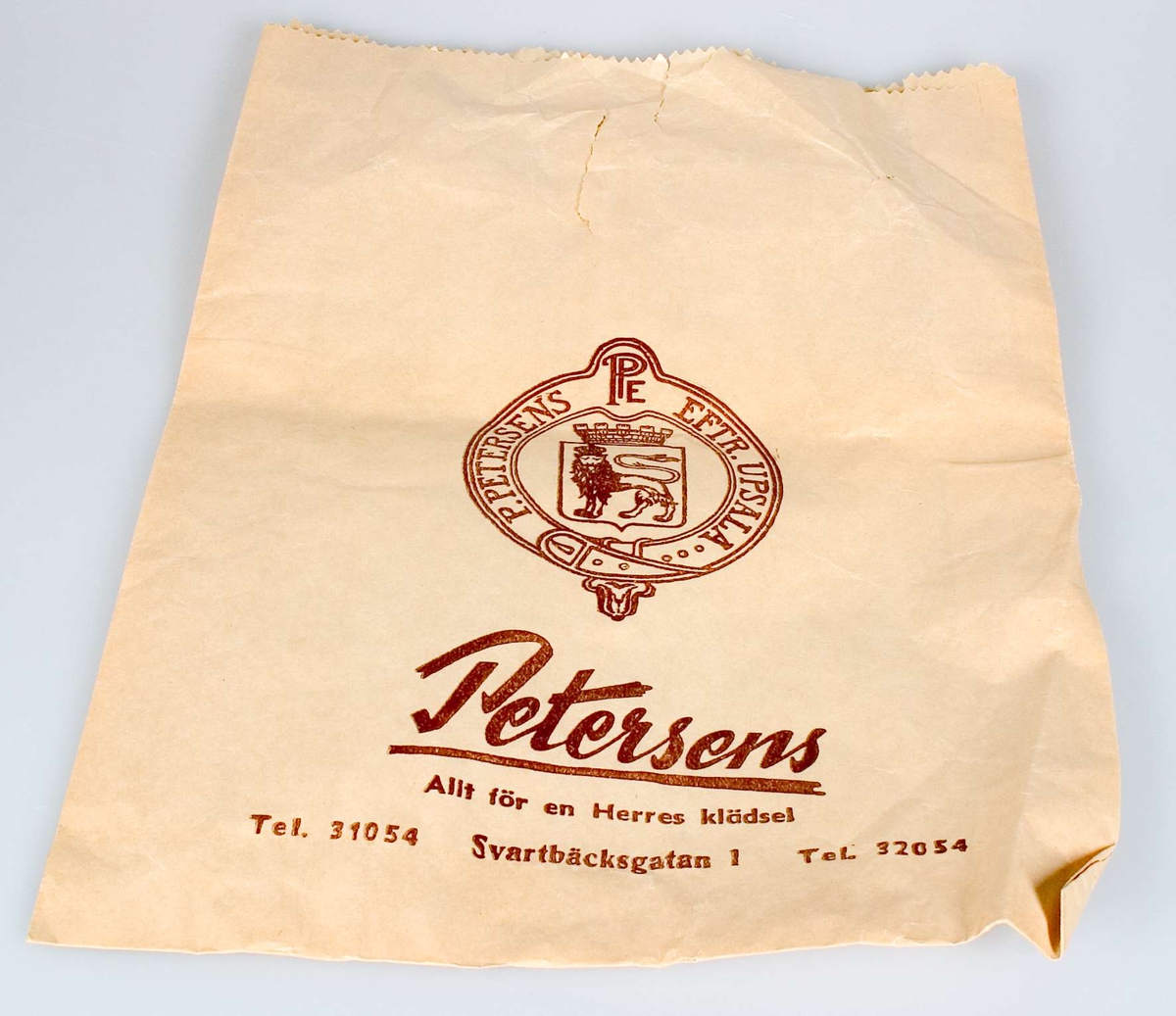 Papperspåse med tryck i brunt: Petersens, Allt för en Herres klädsel, Tel. 31054, Svartbäcksgatan 1, Tel. 32054 samt logotype för P. Peterséns Eftr., Uppsala.