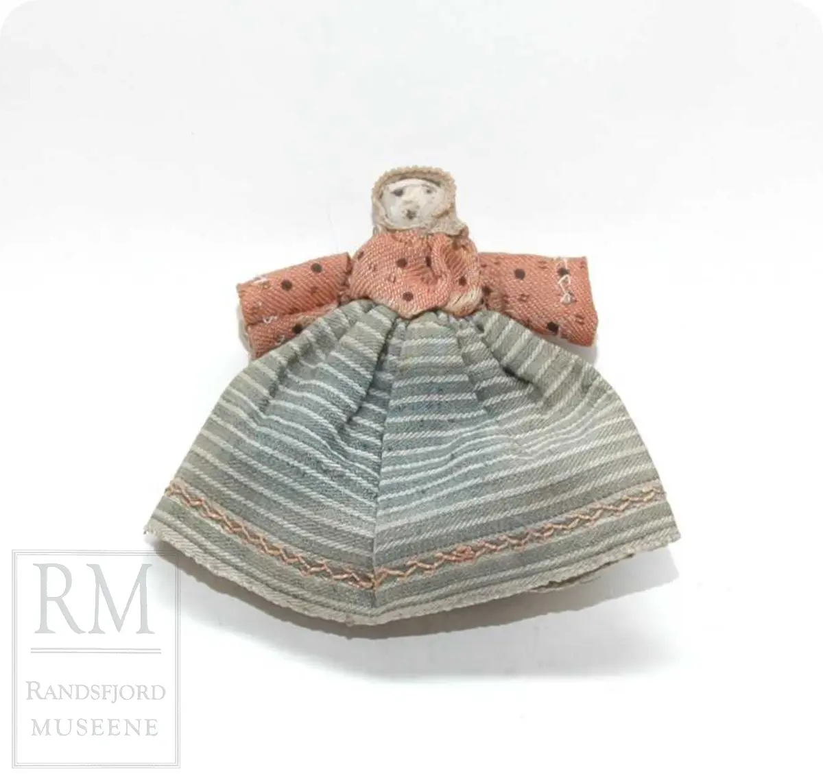 a: Liten dukke i stoff. Flatklemt. To underskjørt, skaut. b. Papir med håndskrift stilet til "Student Nielsen" og merket "Fra en Venninde paa Røros 1865" Tekst: Agtværdige Herr Person! Saasom, dasom og eftersom jeg i disse Dage er berøvet mine fordums gode Talegaver maa jeg i dets Sted gribe til Pennen, for paa den Maade, at faa sige Dem, hvad som egentlig ligger mig paa Hjerted. Det er blit mig fortalt, at De om nogle Dage, agtes at drage ned til Hovedstaden. Den samme Tour staar ogsaa for mig, men at jeg alene kan begive mig afsted skjønner De nok er en Umulighed. Derfor kjære Student, er det min inderlige Bøn til dem, om jeg kan faa slaa Følge med. Jeg ser dem an for at være en ret paalidelig og god Reisefælle, og vover jeg da at gjøre Dem dette Spørgsmaal. Haaber da at De unskylder min Nærhed, slutter jeg disse Linier. Det er mig meget om at gjøre at jeg allerede i Dag kunne faa et par Ord til Svar. Modtag den bedste Hilsen fra Deres hengivne og forbundne Simonette"