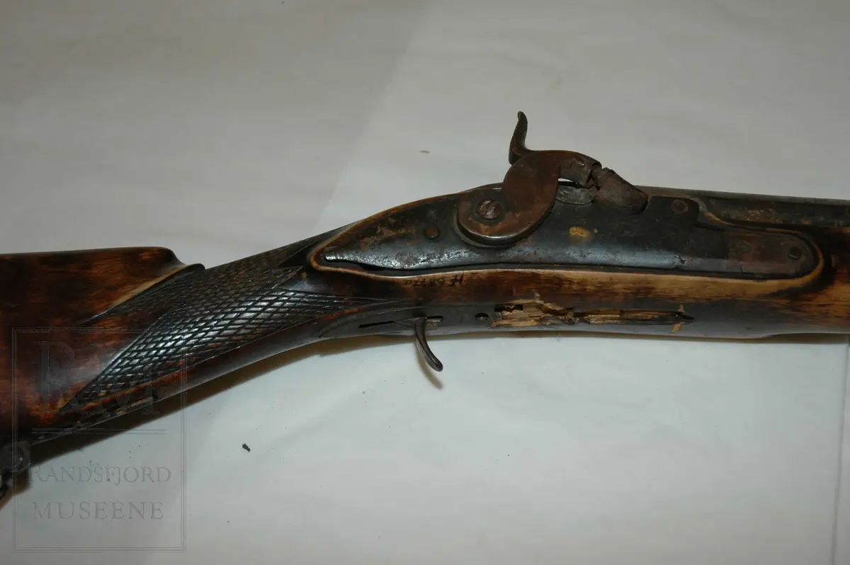 Munnladning, glattløpet m/ perkusjonslås. Skjefte av tre, brunbeiset. Kolben har kinntre og utskjæringer (nettskjæring) på kolbehalsen. Pipe og lås av jern m/ fram og baksikte i messing. To rembøyler med skruer. Ladestokk (b)  av tre er festet under forskjeftet. Bøylebeslag, avtrekkerbøyle mangler. gjennomgående skrue mellom beslag bak pipe og avtrekkerbeslag mangler. Forskjeftet går helt fram til pipemunningen. Har ikke bajonett. Pipen er rund framme og åttekantet bak. Grovkalibret.