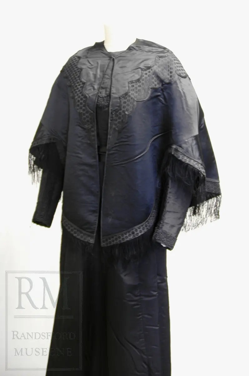 Håndsydd. Svart silkesatin med glanset bakside. Alpakkarenning (?) m / innslag i natursilke. Applikasjon av svart mønstret silkerips. To agramanrosetter med svarte glassperler. Sammensydd i rygg. Lukkes med hekte og malje ved halsåpning foran. Frynser.