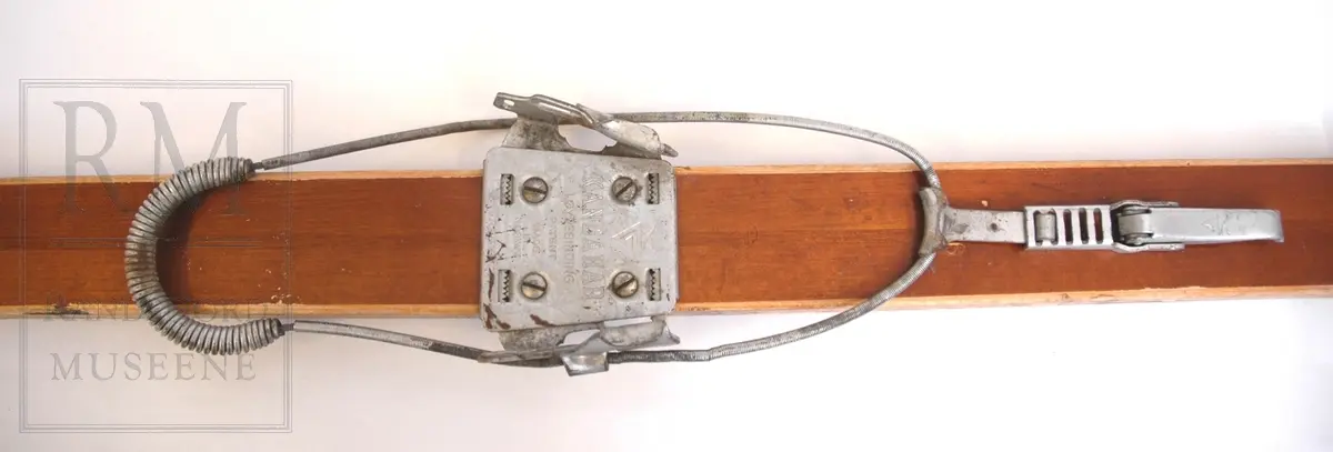 Brunlakkerte treski med fjørbindinger ("Kandahar løypebinding") der det går en fjær bak hælen. Skiene er norskprodusert og av merket Jarl. Varemerket er på begge tupper- en viking i profil med brynje og norske farger.