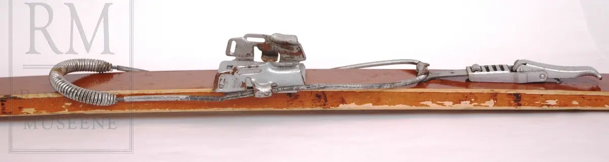 Brunlakkerte treski med fjørbindinger ("Kandahar løypebinding") der det går en fjær bak hælen. Skiene er norskprodusert og av merket Jarl. Varemerket er på begge tupper- en viking i profil med brynje og norske farger.