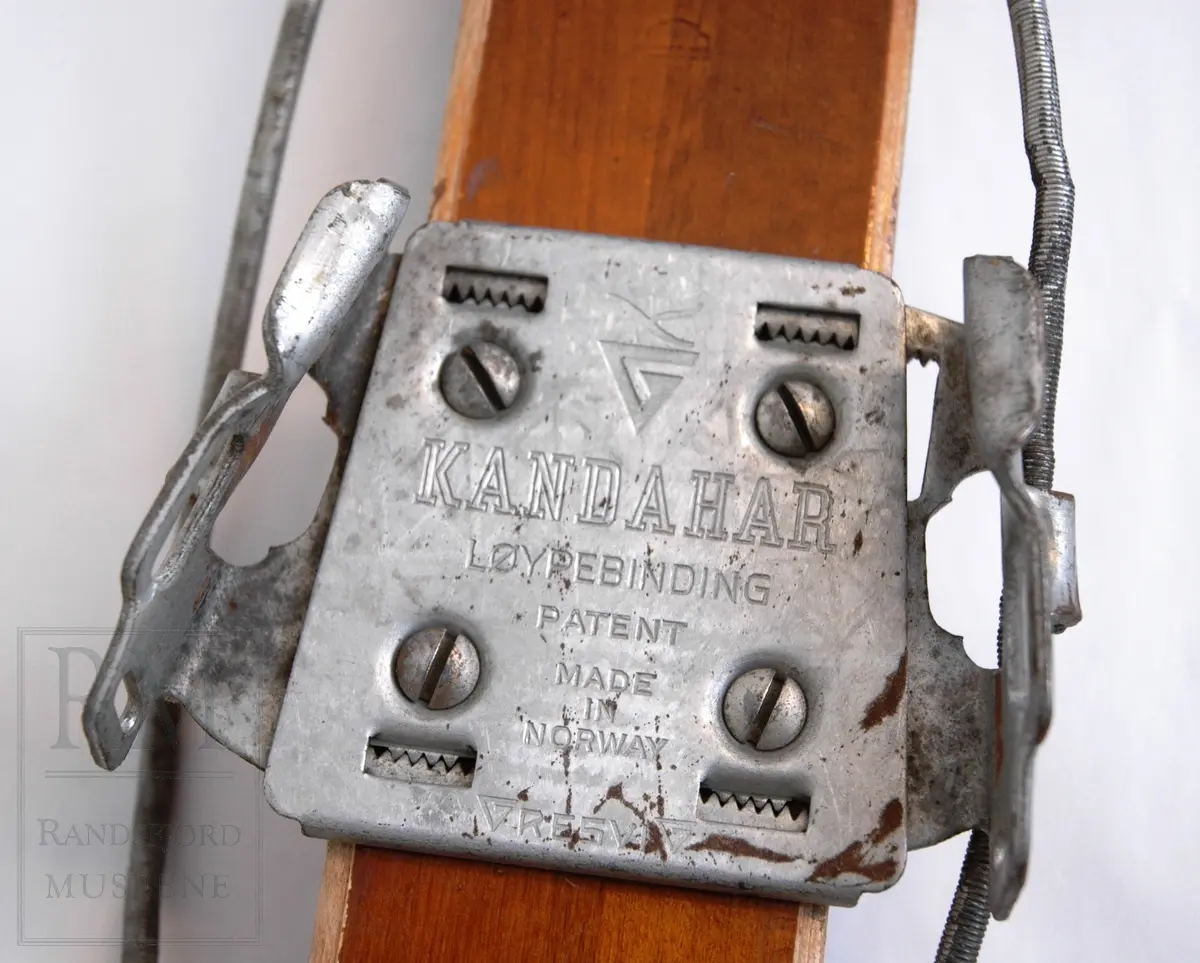 Brunlakkerte treski med fjørbindinger ("Kandahar løypebinding") der det går en fjær bak hælen. Skiene er norskprodusert og av merket Jarl. Varemerket er på begge tupper- en viking i profil med brynje og norske farger.