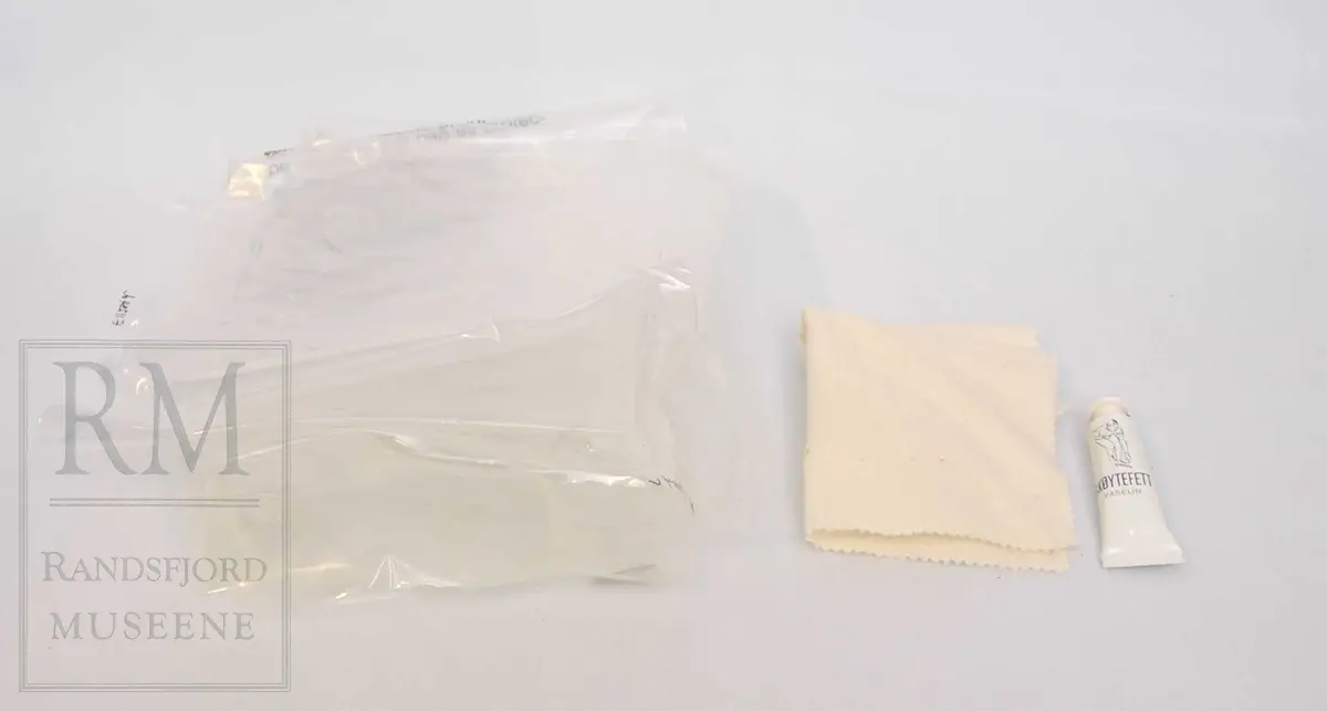 a-b) Skøyter: av svart skinn m / pløse og kant av beige skinn.Røret på skøytejernet avsluttet med plast i begge ender. 
c) plastpose til skøytene.
d) plaststykke.
e-f) Pappeske m/lokk. Brun papp med svart trykk på lokket.
g) pusseklut)
h) skøytefett i tube.