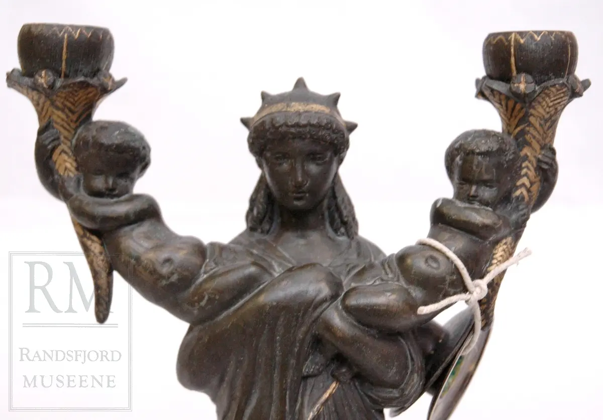 Kvinnefigur på sokkel, med ett barn i hver arm. Barna holder hvert sitt horn (?) med lyseholder. Noe forgylling. Løse lysmansjetter. b) mangler sokkel.
