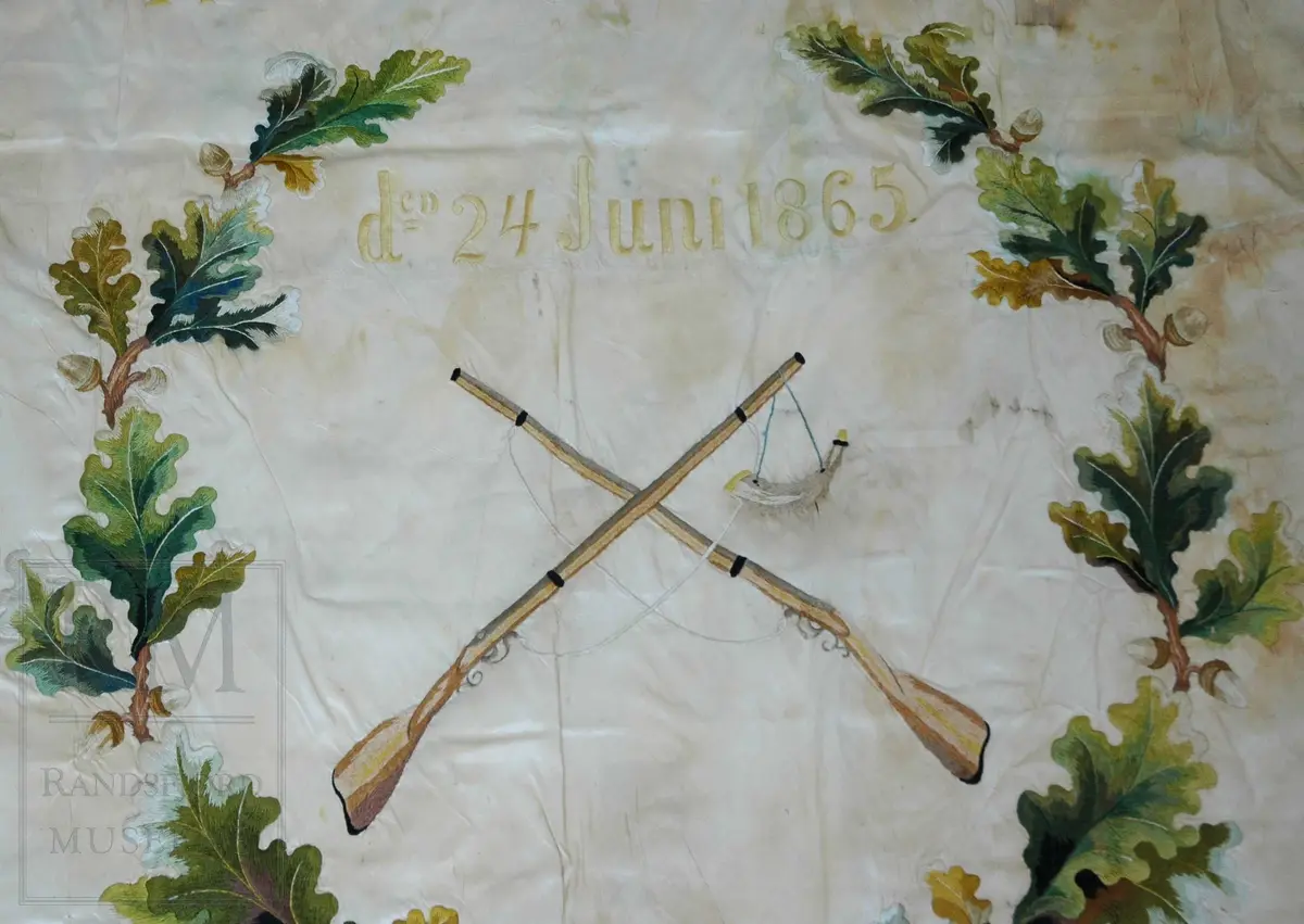 Fane av hvit silke. Løpegang på den ene siden har revnet. Brodert tekst, samt eikebladskrans med sløyfe omkring to gevær med påhengende krutthorn.