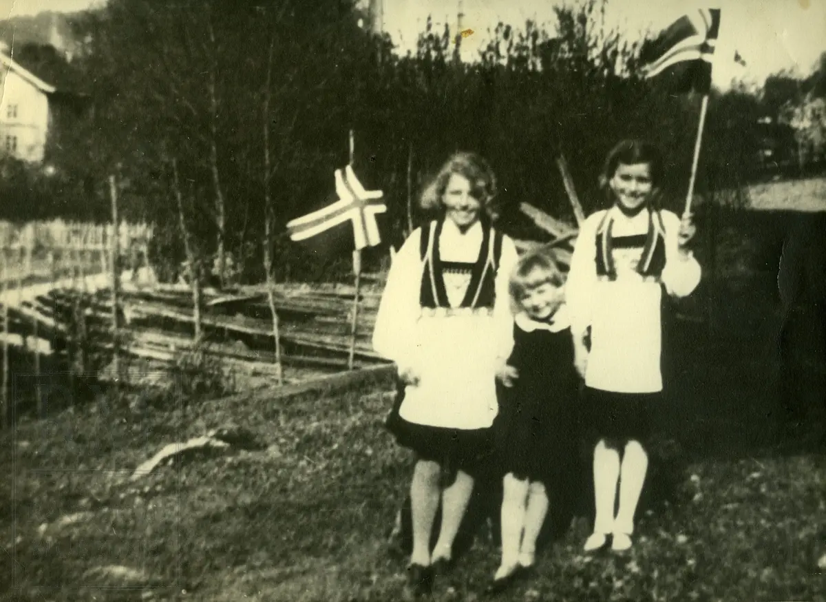 Tre jenter, søstre, med flagg på 17. mai, oppstilt foran en skigard.