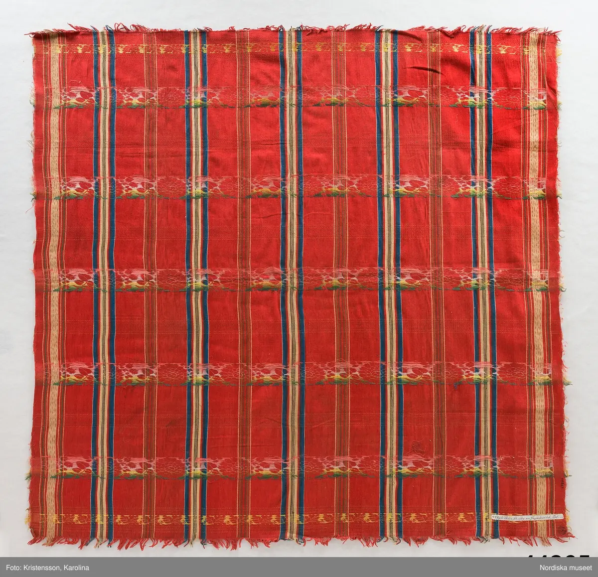 Kvadratiskt halskläde av  halvsiden.  Storrutig  på klarröd botten i satin  med  ränder i schatterande blått, beige, rött och brunt och med jacquardvävda tvärgående blombårder  i rosa, grönt  och gult silke med smala gula  kantbårder. Runtom upprispad kort frans. I ena hörnet en rund svart stämpel med svårläst text: Stockh. Det är Stockholms hallstämpel jfr. likadan t halskläde 44.265 som har en tydligare stämpel  från 1830-talet.
I mycket gott skick.
Troligen en brudschal. Se samma mönster i mörkblått 91.624 från Unnaryd i Småland.
/Berit Eldvik 2012-01-27