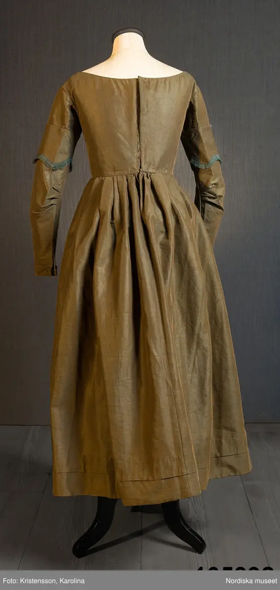 Hel klänning i 1830-talets modesnitt, av fint halvylletyg i kypert med varp av ljusbrunt bomullsgarn och inslag av grönt tunt glansigt redgarn som ger en grönskimrande yta. Livet går ned nästan till midjehöjd, figursytt med 2 framstycken med bakåtflyttade sidsömmar, ett ryggstycke med 6 inprovningar fördelade i två grupper. Vid båtringad halsringning, knäppning med tätt sittande hakar och hyskor. Isydda långa ärmar svängda med 2 sömmar  med 3 veck vid armbågen. Ärmsprund knäppt med hyska och hake samt 4 cm uppslag med rundade hörn. Upptill på ärmen besättning av tyget med stora rundade uddar kantade med klargrön bomullsfrans.  Passepoaler av tyget i alla sömmar. Livet fodrat med skotskrutig bomullslärft i rött, grönt, gult och vitt och i ärmarna ett smalrandigt bomullstyg i blått och rosa. Kjolen sydd av  5 våder, lagda veck runtom i midjan. sprund mitt fram. Uppsytt veck  en decimeter från fållen. Skoning med brun bomullslärft. Handsydd.
Har burits 1838 som ungmorsklänning, alltså av bruden dagen efter bröllopet, av Maria Margareta Mårtensdotter från Kysings, Vall sn på Gotland. Möjligen tillsammans med sidenhalsklädet 105385.
Anm. I  gott skick.
OBS Klänningen sitter bakochfram på fotona, knäppningen är fram.
/Berit Eldvik 2011-06-13