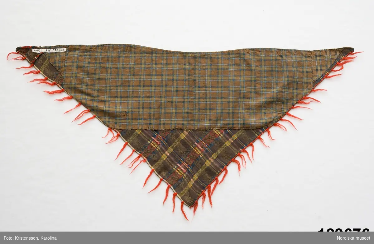 Trekantigt halskläde av troligen jacquardmönstrad bomullssatin.  Grönbrun botten randig i  ljusare grönt, gult och blått med invävd bladslinga i bottenfärgen. Tvärgående mönsterbårder i rött och blått  samt smalare i gult och grönt, vilket ger ett rutigt  tyg. Tvärbårderna i inslagsflotteringar.   Tyget har 2 materialskarvar. I ytterkanterna iknuten gles frans av rött ullgarn.Fodrad till större delen med ett grön/brunrutigt bomullstyg, snibben bak ofodrad.
 Tillverkad av delar från ett kvadratiskt halskläde.
/Berit Eldvik 2012-02-09