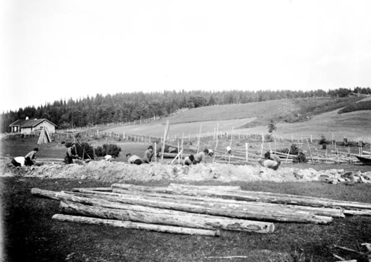 Graving av av tømmerkanal i Sørum i Akershus, antakelig i nærheten av Bingen lense.  Fotografiet er tatt på et engareal der ti karer med spader var i ferd med å grave ei grøft over ei eng, med sikte på å opparbeide en liten kanal der de kunne trekke tømmer på vannspeilet.  Her ser vi ikke sjølve kanalen, men massene karene hadde kastet opp fra den forsenkningen de grov.  I forgrunnen lå det en del barket, øyet fløtingstømmer på grasbakken.  I bakgrunnen ser vi et kulturlandskap med eng- og ækerarealer, og på en bakkekam til venstre et lite husmannsbruk.  Til høyre i bildet ser vi en av stevnene på en robåt av den typen fløterne i denne delen av Glomma brukte. 