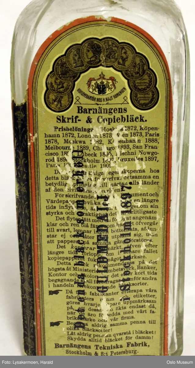 Flasken har etikett over tre sider. Den har mørk beige bunnfarge, og trykk i sort, blått, rødt og gull. Tegning av en bjørn som rører i en bøtte med blekk under teksten "FABRIKSMÄRKE".