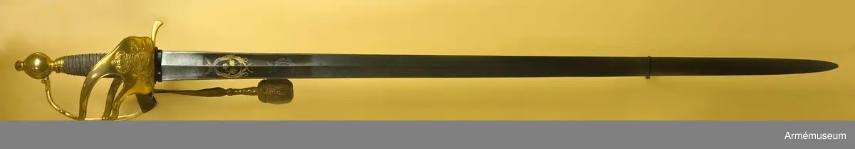 Fäste av förgylld mässing med två yttre och en inre sidobygel. Parerplåten har en krönt sköld omgiven av stiliserade palmkvistar. Kaveln klädd med hajskinn och lindad med en tvinnad,  förgylld mässingtråd. 

Klingan är rak och tveeggad. Bredd vid fästet: 37 mm. Upptill rester av blånering och dekorerad med tre kronor, omslutna av två palmkvistar.