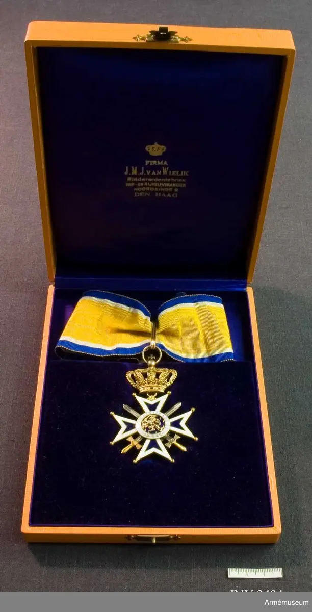 Ask till Nederländsk Oranien-Nassau orden. Utförd i apelsinfärgat klot med på lockets översida två  korslagda svärd och däröver riksvapnet med på banderollen texten: JE MAINTIENDRAI. (= Jag vill hålla ut.) I locket lila siden med text i guldbokstäver: Firma J M J van Wielik  Riddererdenfabriek Hof-en rijksleverancier Noordeinde 9 DEN  HAAG. Botten är sammetsklädd i lila och urtag finns för orden (fördjupning).