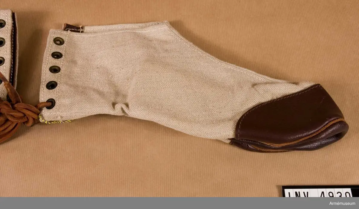 Ladda. Stl 43-45. Av beigefärgad smärting med brun sula/tåskoning. Stänges mitt bak med öljetter, genomdragna med lädersnören. Skyddsbeklädnad i vinterterräng till snödräkt. (M 7336-291000).