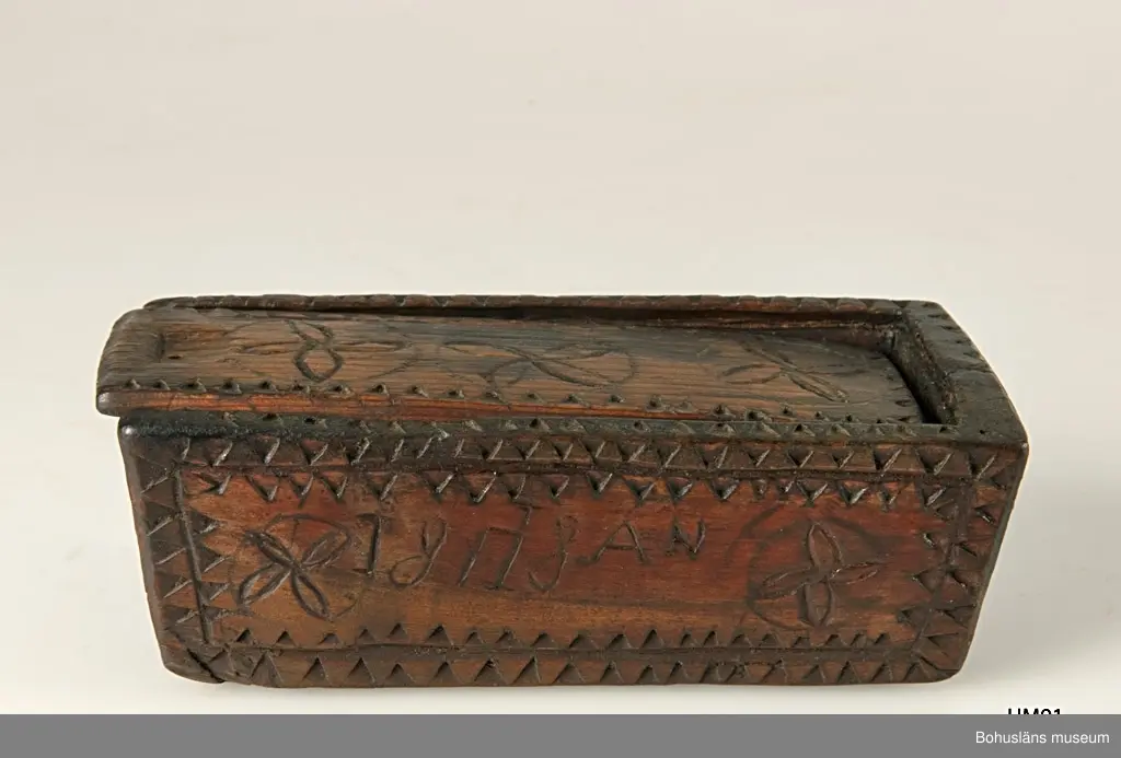 Rektangulär låda med skjutlock och något snedställda kortsidor.
Skuren dekor bestående av rosetter, kors samt uddsnitt längs kanterna.
På en av långsidorna är årtalet "1848" och initialerna "AN" inskurna.
Inuti ligger en äldre etikett från ca sekelskiftet.
Kan vara en friargåva.

Litteratur:
Gjaerder, Per, Esker og tiner, C. Huitfeldts förlag, Oslo, 1981, s. 41.
Knutsson, Johan, Friargåvor, Nordiska museets förlag, Stockholm, 1996.

Ur handskrivna katalogen 1957-1958:
"Trälåda
1870-1887
Bottenmått: 14 x 5,5 H. 5. Trä med skuren ornering. Föremålet helt. Bohusl."

Lappkatalog: 84