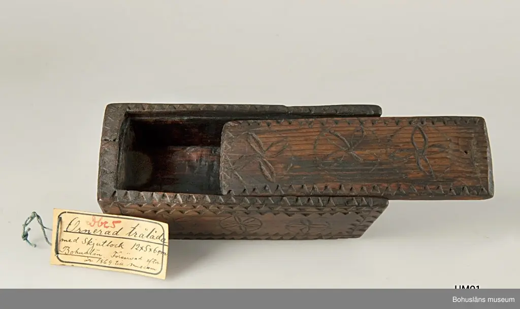 Rektangulär låda med skjutlock och något snedställda kortsidor.
Skuren dekor bestående av rosetter, kors samt uddsnitt längs kanterna.
På en av långsidorna är årtalet "1848" och initialerna "AN" inskurna.
Inuti ligger en äldre etikett från ca sekelskiftet.
Kan vara en friargåva.

Litteratur:
Gjaerder, Per, Esker og tiner, C. Huitfeldts förlag, Oslo, 1981, s. 41.
Knutsson, Johan, Friargåvor, Nordiska museets förlag, Stockholm, 1996.

Ur handskrivna katalogen 1957-1958:
"Trälåda
1870-1887
Bottenmått: 14 x 5,5 H. 5. Trä med skuren ornering. Föremålet helt. Bohusl."

Lappkatalog: 84