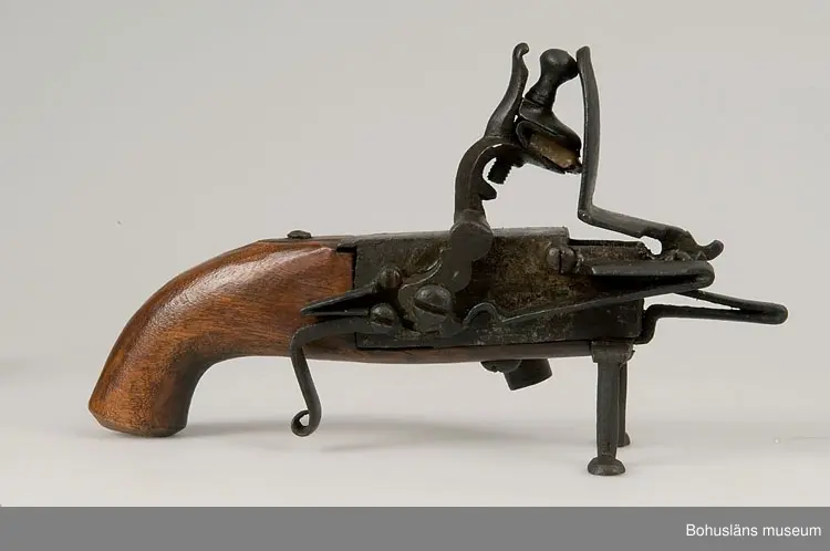 Ur handskrivna katalogen 1957-1958:
Elddon, "1800".
L. 22, H. c:a 13 cm; ett flintlås monterat m. handtag av trä och 2 fötter av järn; på sidan en lucka på gångjärn, m. regel, på denna (d.v.s, på luckan) en ljushållare av järn. Helt.

Lappkatalog: 90