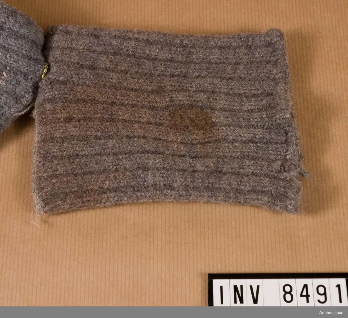 Samhörande i par 8491-2.
Stickad i grått yllegarn, två aviga och två räta maskor. Stickad som en mudd som sytts ihop ett stycke in i nederkanten, så att det bildats ett hål för tummen. På så vis täcker mudden halva handen och hålls samtidigt på plats.