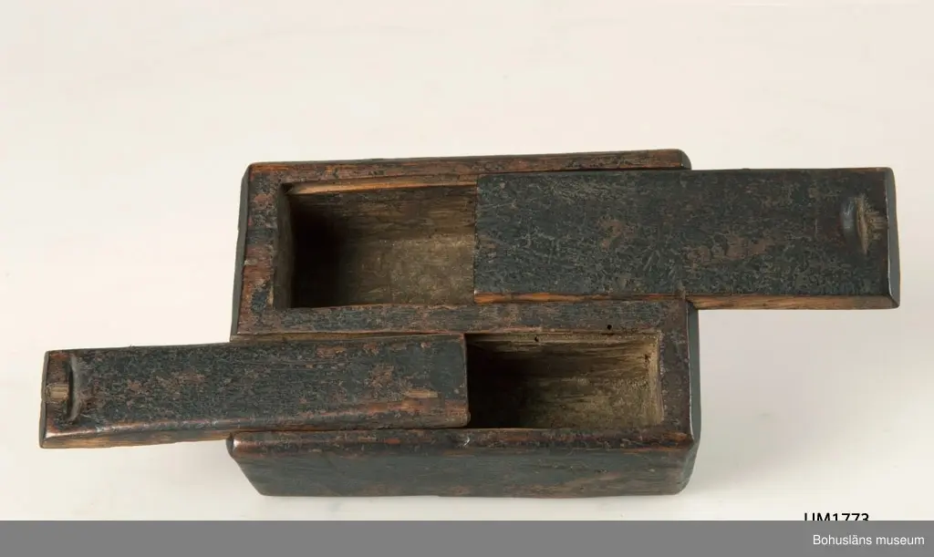 Rektangulär låda med två skjutlock, vilka dras ut åt varsitt håll.
I det större facket förvarades en flintbit eller ett eldstål och i det mindre fnösket att fånga gnistan med.
Den svartmålade färgen har flagnat runtom.
Något skadedjursangrepp.

Litt.; Gjaerder, Per, Esker og tiner, C. Huitfeldts förlag, Oslo, 1981, s. 48-50.

Ur handskrivna katalogen 1957-1958:
Ask, dubbelt skjutlock
Bottenmått. 12,5 x 8 H: 4
Trä, brunmålat. Föremålet helt. Forshälla, Bohusl.

Lappkatalog: 84