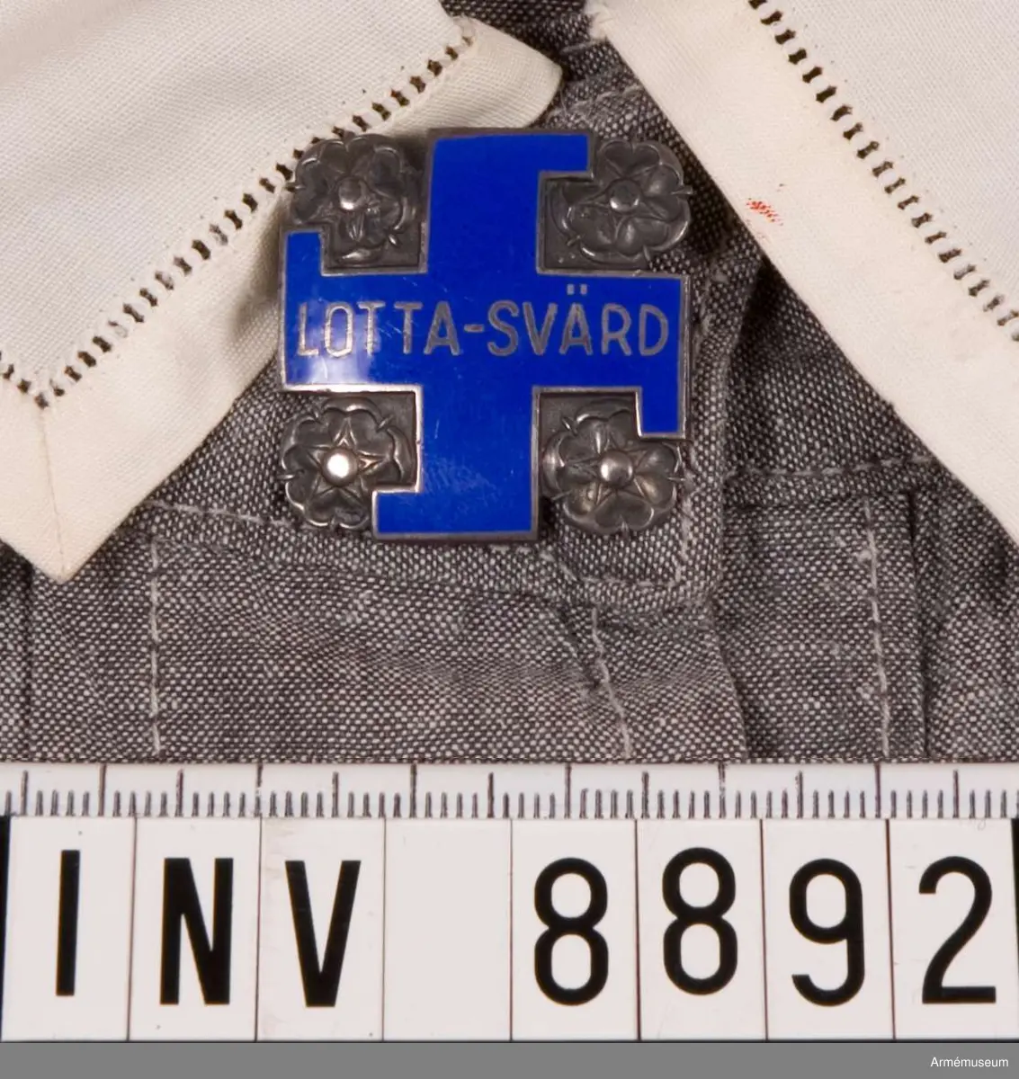 Av silver. Visar ett svastikakors i blå emalj med silverkant och namnet LOTTA-SVÄRD. Korset är omgivet av rosor i silver. Frånsidan bär silverstämplar och präglat nummer 63404.