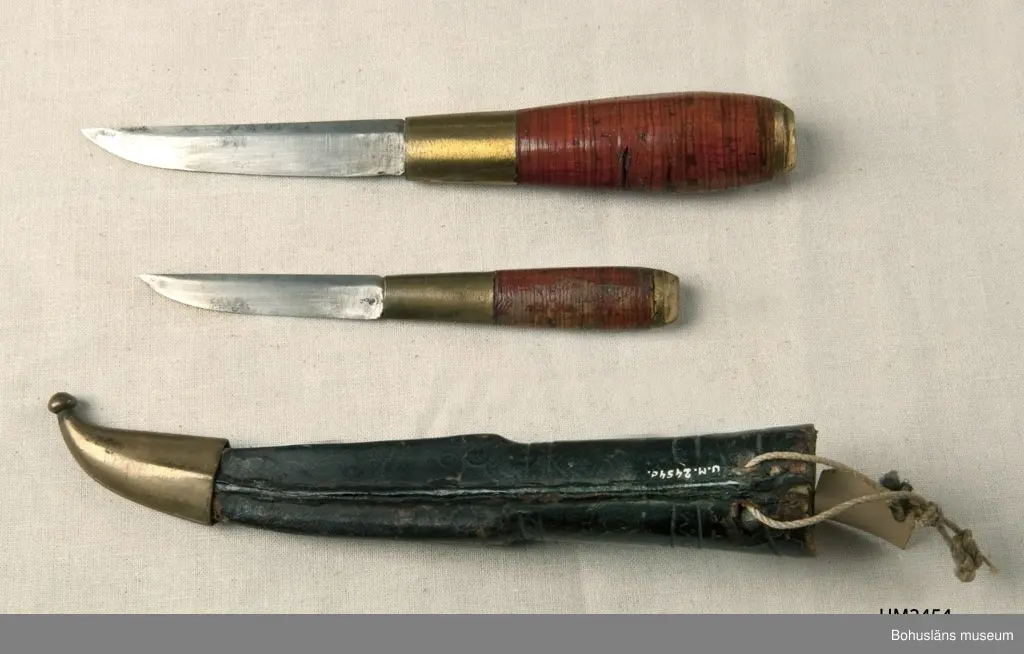 Två knivar i en och samma slida. En större (längd 16 cm) och en mindre (längd 12 cm).
Knivarnas skaft är gjorda av näver som packats tätt kring tången.
Lädret i slidan har pressat mönster och har färgats svart. Mässingstoppen har ristad/graverad dekor. Slidan är fodrad med ett ljust gröntonat papper.

Knivarna är tillverkade i Kalajoki, en stad i Norra Österbotten i Finland. Slidans doppsko är tillverkad i en s.k. Kauhava-modell. Kauhava är en stad i Södra Österbotten .

Ur handskrivna katalogen 1957-1958:
Två slidknivar i en slida
Kniven a) L. 16 cm; m. skaft av mässing och läder; kniven b) L. 11,8 cm; lik föreg. Slidan c) L. 16 cm; svart läder, skoning av mäss. Föremålen hela.

Lappkatalog: 29