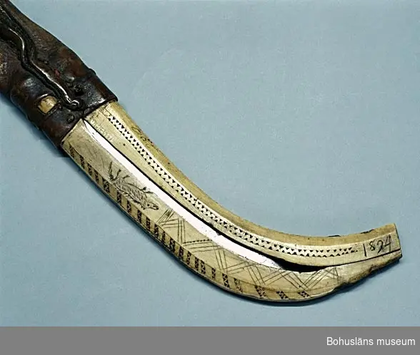 Mått ovan avser kniv och slida tillsammans.

Hemsmitt kraftigt knivblad med enkelt skaft av masurbjörk. Knivbladet 22,4 cm långt och något bredare vid änden, 5 mm tjockt.
Hela kniven 32,5 cm lång.
Slidan av renhorn, övre skaft troligen av barkgarvat renskinn med kraftig upphängningsögla av råhud. Slidan 30 cm lång, tjocklek 15 mm.
Renhornet tillverkat av två svängda delar bildande en nedre böj och med ett mellanrum, en slits, på var sida där knivens platta sidor syns igenom. Dekorerat längs sidor och kanter med ristade bårder av vågor och sicksacklinjer samt en ren och tre vargar (hundar?). På ena platta sidan skrivet med brun färg "Aat" (med en uppåtvänd halvcirkel över a) samt i toppänden inristat "1894" . Den ristade dekoren är ifylld med mörkbrunt färgämne. Skaftet sammansytt med sentråd och lintråd.

"A O T 1894" kan avse köparen och är särskilt ingraverat.

Lappkatalog: 7