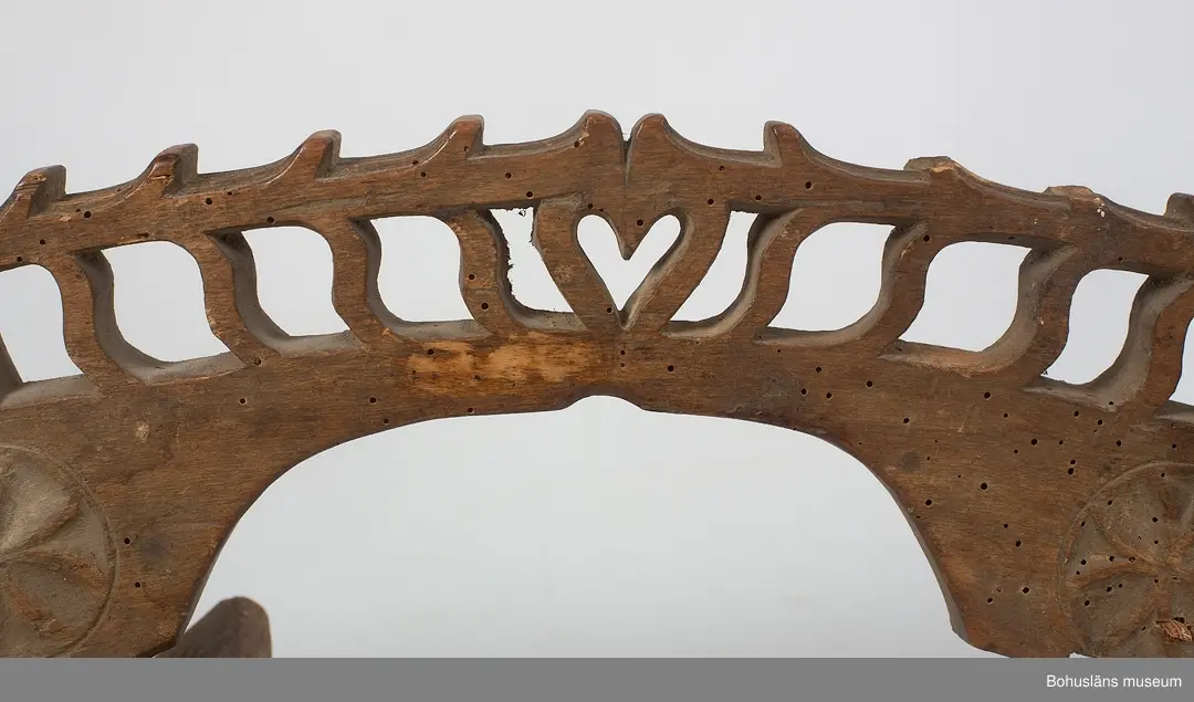 Böjd, genombruten båge med karvsnittsdekor på båda sidor bestående av rosetter. Längst ut på vardera ända är ett drakhuvud.
Rektangulära fästen, vilka vilar på hästens manke. Karvsnittsdekor med rosetter på en av dem. Den andra är sekundär och har skurna cirklar samt är defekt.
Ett drakhuvud är sekundärt.
Skadedjursangrepp.

Litteratur: Edvardsson, Ingmar, Rack, ryss och rissla, Stockholm, 1995, 
s. 66 samt bild s. 42.
Kjellberg, Sven T.: Svenska selkrokar av trä i Nordiska museet. Fataburen 1926. 
Nylén, Anna-Maja, Hemslöjd, Håkan Ohlssons förlag, Lund, 1978, s. 396-401.

Ur handskrivna katalogen 1957-1958:
Selranke av trä
I 3 delar a-c, a) = oket L. 59 cm; b) "stödplatta", L. 19,5 cm; c) d:o, L. 20 cm (nygjord). Oket har snidad dekor i genombrutet arbete m. drakhuvud i ändarna (ett nygjort); hela hela selranken har karvsnittsdekor. Omålad. Maskäten. 

Lappkatalog: 40
Hugo Hallgren var son till salteriidkaren och handelsmannen Johan Jacob Hallgren och Emelia Gerle. Företaget började sälja brissling på 1850-talet och 1864 startade man konservfabrik på Gullholmen som senare döptes till J. J. Hallgren & Söner. Sönerna var Gustav Richard, f. 1858 och Oscar Hugo, f. 1867.  Här introducerades flera nyheter inom ansjovisindustrin,  bl a Sveriges första skinn- och benfria delikatessill. Företaget var också först med att lägga in ansjovis i hermetiskt slutna kärl av bleckplåt. 1898 köptes Hallgrens konservfabrik upp av det nybildade storföretaget AB Sveriges Förenade Konservfabriker (SFK). 1901 flyttade Hugo Hallgren till Ellös där han startade en ny konservfabrik.



För uppgifter om förvärvet, se UM5155.