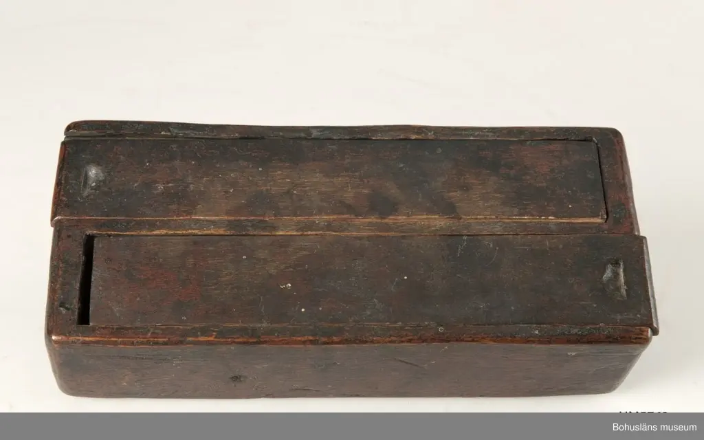 Rektangulär låda, uppdelad i två parallella fack med varsitt skjutlock upptill.
Målat med mörkt röd färg.
Inuti ett av facken finns fragment av fnöske.

Litt.; Gjaerder, Per, Esker og tiner, C. Huitfeldts förlag, Oslo, 1981, s. 48-50.

Ur handskrivna katalogen 1957-1958: 
Trälåda m. två skjutlock
a) L. 22,5 Br. 9,7, H. 5,3 cm, rödmålad; m. 2 fack, under m. ett skjutlock b) och c); deras L. 21,2 cm, Br 4 cm.

Lappkatalog: 84
