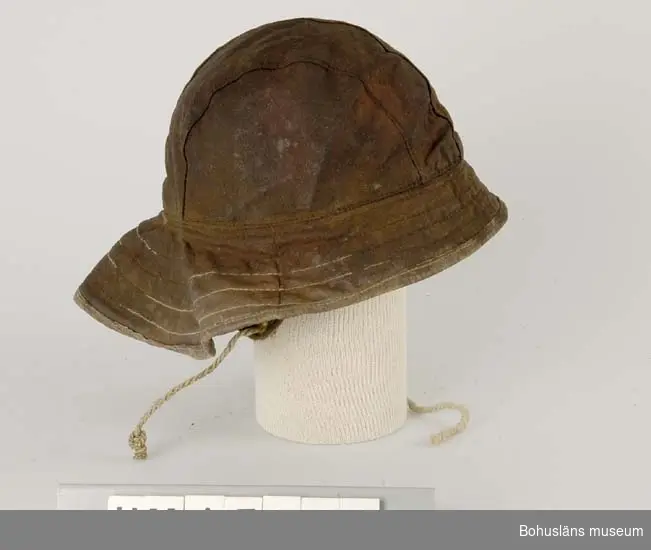 Hatt med kulle sydd i fyra längsgående stycken. Brätte som är större  
bak i nacken än framtill. Foder med öronlappar och ett snöre att knyta
under hakan. Isydd lapp anger storlek 57. Hatten är brun-röd på ut-   
sidan. Fodret är rosa.                                                
Brättet är större bak för att förhindra vatten att rinnna in vid      
nacken.                                                              

Ingår i redskapsbestånd ur sjöbod från Hällsö, Havstenssund, Tanum sn.
Se UM17521.