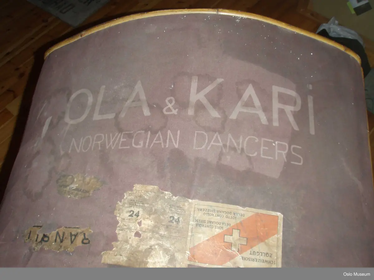 Reisegarderobe, amerikakoffert brukt til ballettutstyr i rotting med buet og lerret-trukket tak. 2 bærehanker. Åpning i front med beslag av metall. Innvendig trukket med ullstoff (ullteppe).