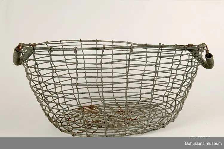 Rund korg vilken är svagt konande nedåt. Två ledade bärhandtag omvirade med ståltråd.
Korroderade.
Mått på den andra korgen: L 53,5, B 55, H 22 cm