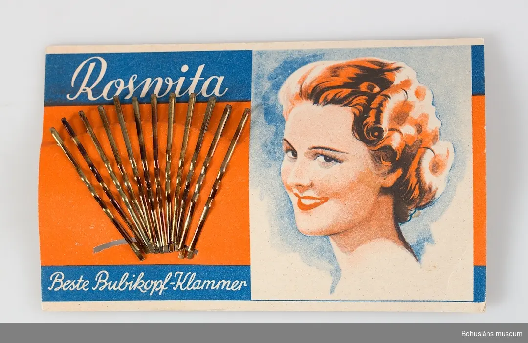 Rektangulär, pappskiva med dekor i rött, blått, vitt och text: "Rosvita. Beste Bubikopf-Klammer." 12 hårnålar av mässing isatta i solfjädersformat. Tre kartor, en skadad.