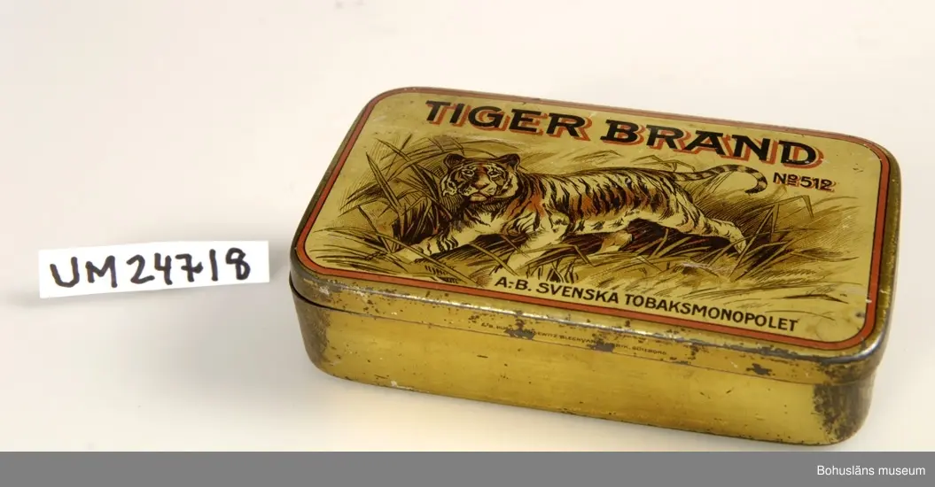 Föremålet visas i basutställningen Kustland,  Bohusläns museum, Uddevalla.
471 Tillverkningstid 1940-TAL ?

Locket sitter fast i ena långsidan. På locket finns det bild på en tiger och text: "TIGER BRAND NO 512 A.-B. SVENSKA TOBAKSMONOPOLET".
Asken är tom.
När asken kom till museet låg det små svenska flaggor inuti. 
Se UM24719.