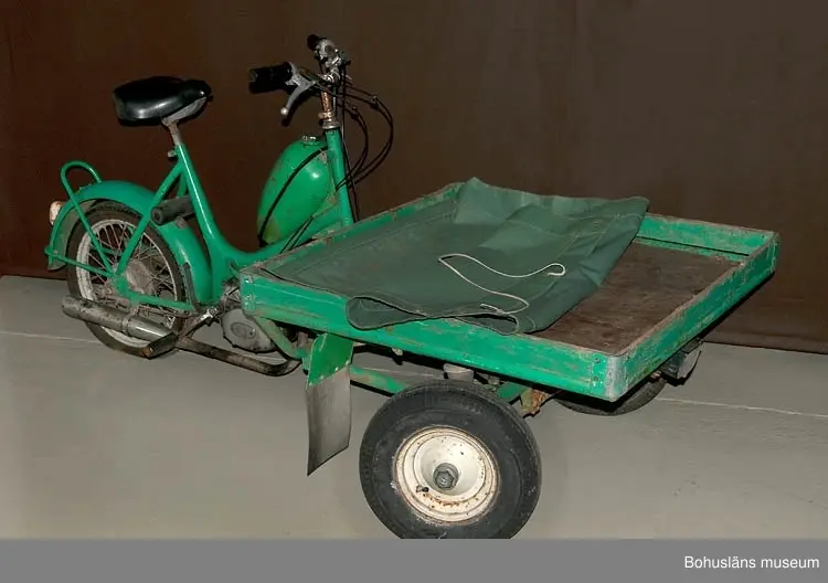 Måttuppgifter flaket: 195 x 195 cm; höjdmåttet avser höjd vid styret.
Grön presenning, fästad i flakets bakre del.
Mopeden är inköpt i Strömstad 1973 och använd på Sydkoster till slutet av 200
Mopeden ingår i insamlingen i projektet Moderna skärgårdsbor, UM027511 - UM027595 och kommer från Sydkoster.