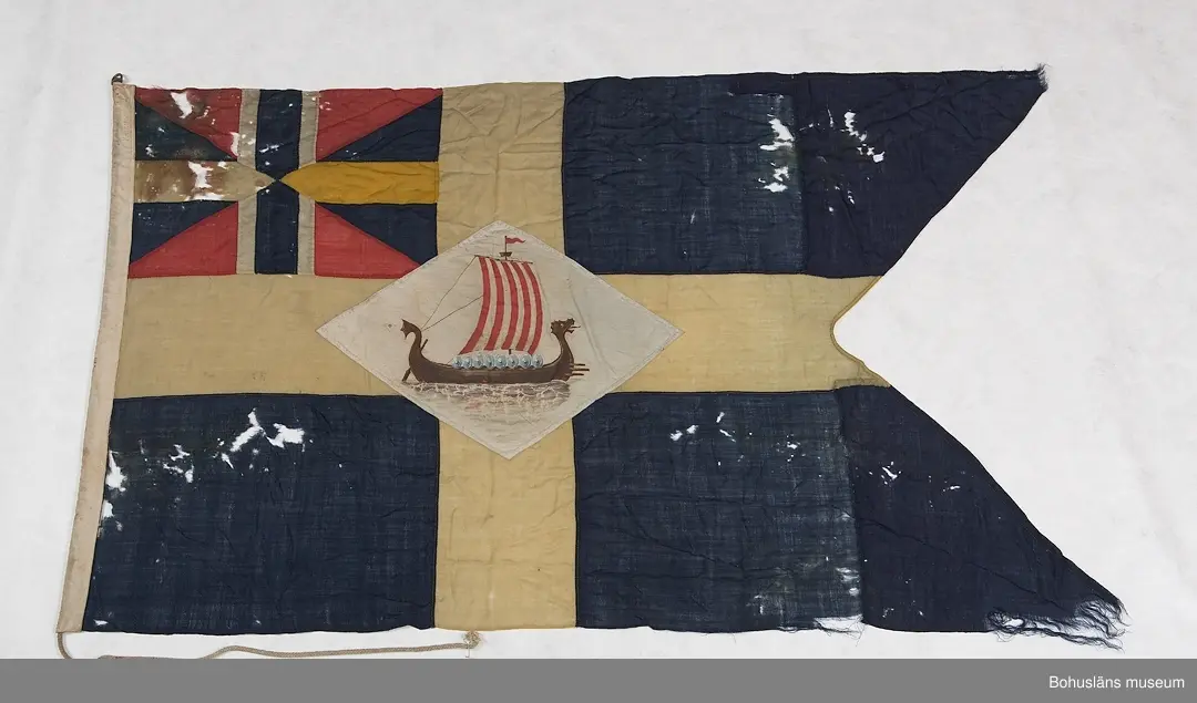 Segelsällskapet Vikens flagga, SS Viken. 
Svensk tvåtungad  flagga med parti "Silllsallaten", svensknorska unionsflaggan i övre hörnet samt infällt handmålat rombiskt emblem placerat mitt i det gula korset. På emblemet är målat ett vikingaskepp. 
Märkt "KASEN" med svart märkbläck. Hand- och maskinsömnad.
Skador. Eventuellt har flaggans form ändrats.