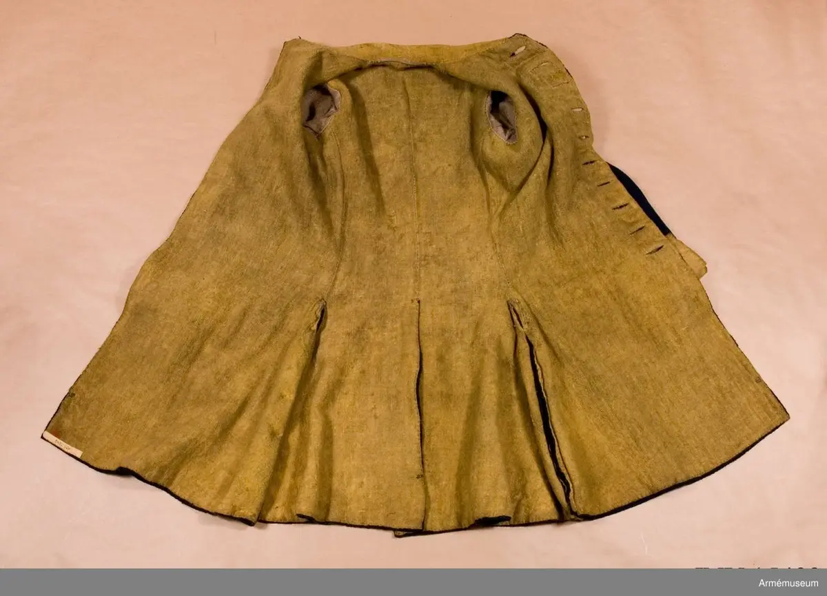 Rock av blått kläde med gul krage och ärmuppslag. Fodrad med gul boj i livet och med linne i ärmarna. Försedd med tennknappar.