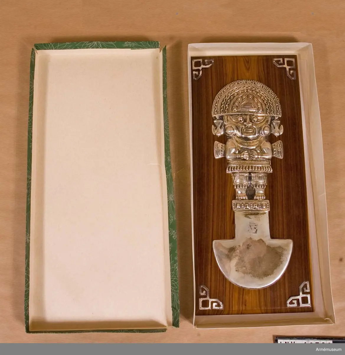 På en brun träplatta i mahogny är monterad en kultfigur från Mexico eller Peru i silverarbete. I plattans hörn sitter ornament i silver. Figuren är märkt "La Plata 325".

Förvaras i pappask, grön med vitt mönster.