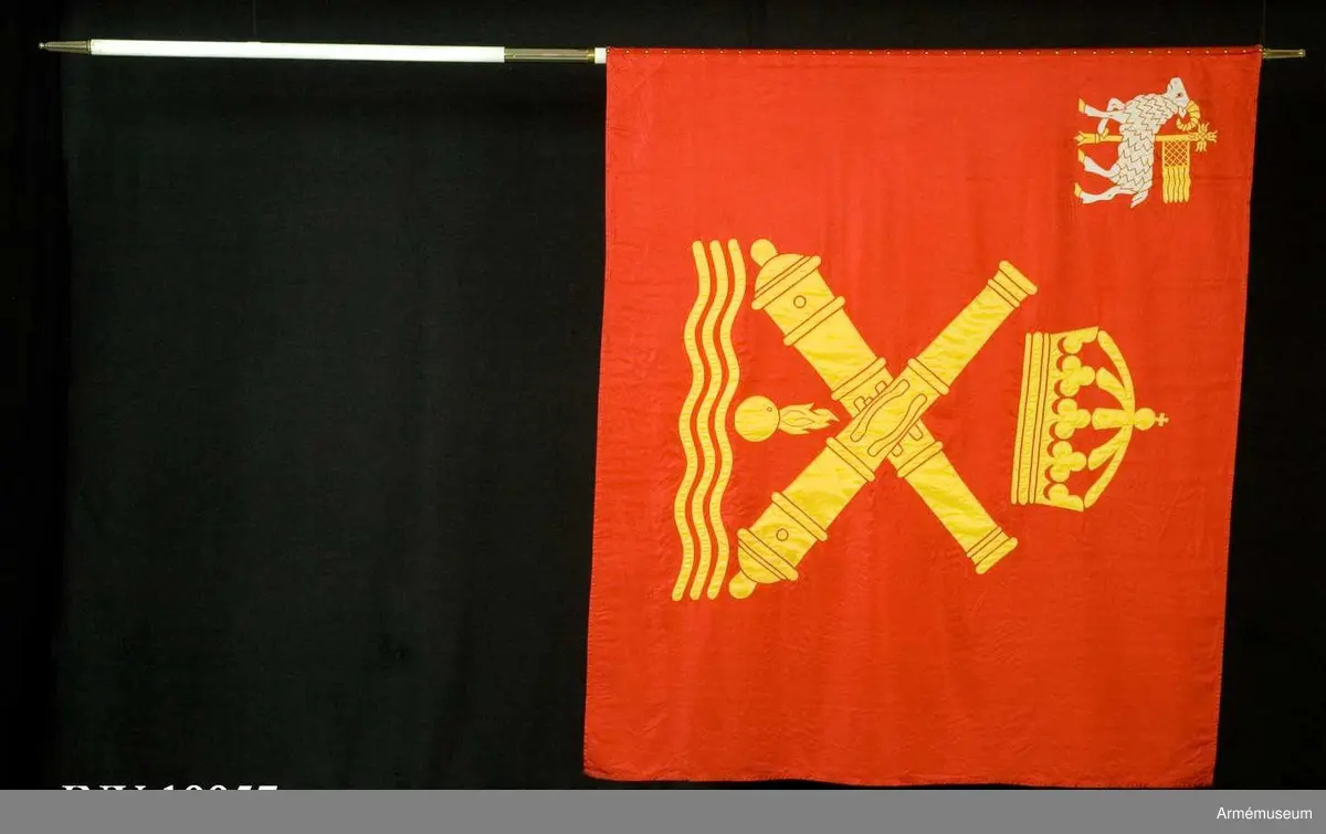 På röd duk i mitten fd kustartilleriets vapenbild; två korslagda eldrör av äldre modell under en kunglig krona och över en flammande granat och vågor, allt i gult. I övre inre hörnet landskapet Gotlands vapenbild; en vit vädur med gul beväring bärande på en korsprydd stång ett banér med bård och flikar, allt i gult.

Applikationerna i gult sydda med maskinsöm i zig-zag. Även vitt fansiden med röd och vit maskinsöm. Fäst vid stången med 36 st tännlikor i rad 2+2 st. Delad vitmålad slät stång.