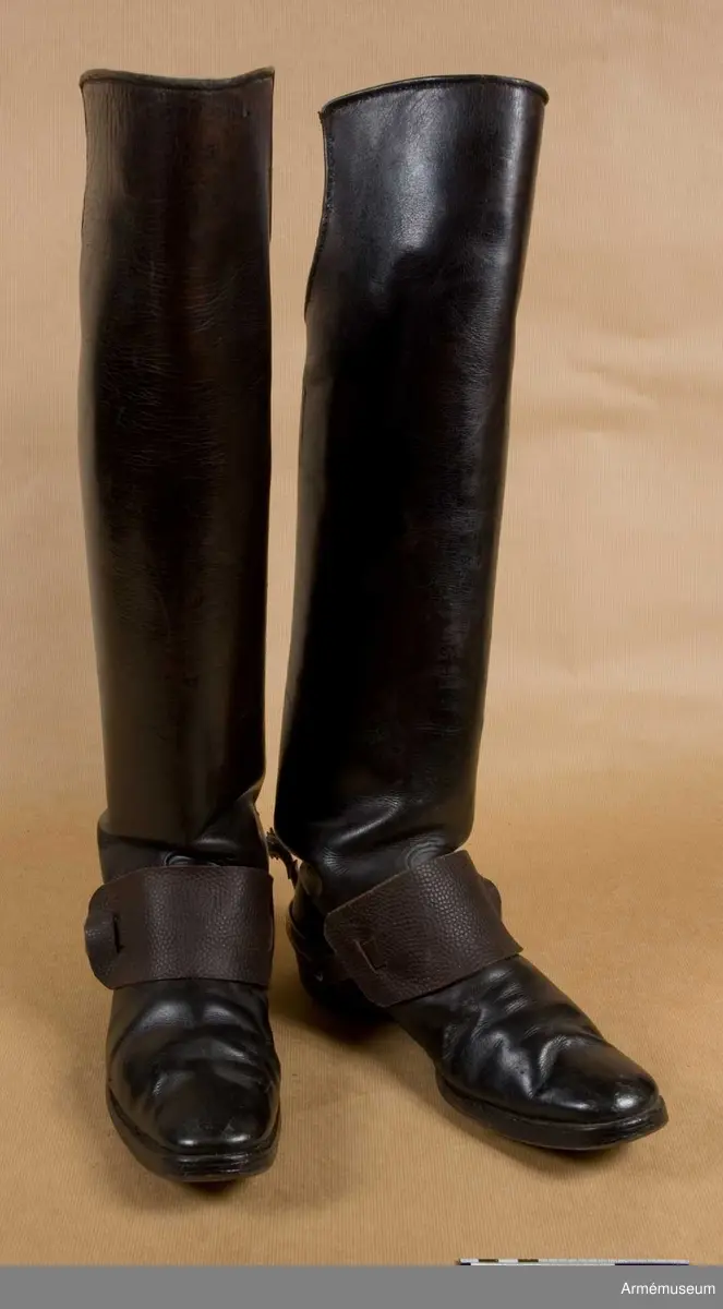 Av svart läder. Raka med en större (ca 15 cm) utskärning för knävecken baktill. På stövlarnas baksida vid klacken finns ett sporrfäste i läder.