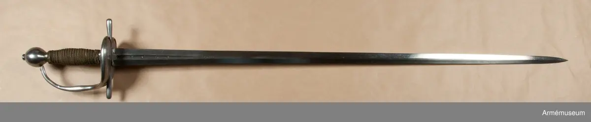 Klingan är tveeggad med flata åsar. Upptill på var sida har den en blodrand, som på yttersidan är 16,5 cm lång. Under den nedra ändan på varje blodrand finnes ett av prickar bestående onament.

Fästet är av polerat järn och av drabanttyp. Knappen är svagt ellipsiodisk med rygg fram och bak. Diam. sidled är 3,95 cm, men över ryggarna 4,15 cm. Höjden är 4,3 cm. Nitknappen är låg och närmast cylindrisk. Halsen är ävenledes låg med konisk. Höjden av hals, knapp och nitknapp är sammanlagt 5,25 cm. 
Kaveln är spolformig med flat rand fram och bak, men sidorna är avrundade. Upp- och nedtill omgives den av en ringliknande, platt flätning, "krans", av rätt varierande bredd, i huvudsaken dock 0,7 cm, vilken flätning består av mässingstråd, men är hopsnodd av två parter. I övrigt är kaveln lindad med 4 mässingstrådar, av vilka två är rätt grova, den ena höger- den andra vänstersnodd, men de båda övriga rätt fina.
Parerstången är nästan rak och blir mot ändarna litet bredare. Den främre änden är avrundad, men den bakre har mer tvär avslutning. Armarna ha spetsovalt tvärsnitt. Den största bredden är för den främre armen 1,1 cm, men för den bakre 1,25 cm. Den största tjockleken är för bägge armarna 0,85 cm. Avståndet mellan den främre eggen och parerstångens främre ändan är 6,8 cm, men mellan den bakre eggen och den bakre änden 5,9 cm. 
Parerplåten är hjärtformig och har förhöjda kanter, vilka sistnämda egentligen bestå av spänger, en på var sida, vilka från parerstångens bakre arm gå till handbygelns nedre del. Tomrummen mellan spängerna och parerstången, resp. handbygelns nedre del, ha utfyllts med ett par kupiga plåtar, som faslötts med mässing. Parerplåten är något större på ytter- än på insidan. Avståndet mellan klingans mitt och parerplåtens kant är på det förstnämda stället 4,6 cm, men på det sistnämda 4,5 cm. 
Handbygeln utgår från korset, har spetsovalt tvärsnitt och den största bredden litet ovanför parerplåtens spets. Dess övre ända är instucken i ett hål i knappen. På handbygelns innersida nära övre ändan är bokstäverna AD inslagna. Tumbygel finnes icke.