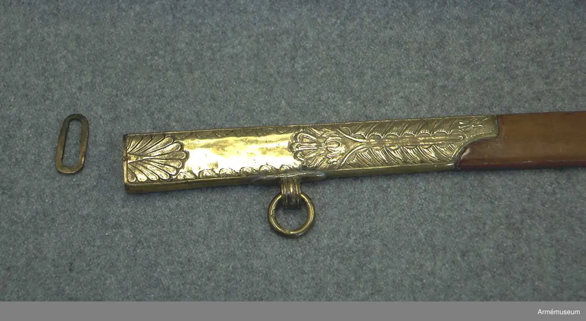 Balja med beslag av förgylld mässing, mittparti läderklätt. Släpsko av stål. Baljan är av brunt läder och har munbleck och mycket lång släpsko av förgylld mässing, som är driven i stiliserade blomornament m.m. 
Släpskon är längst ned på änden och vid framsidan skodd med stål. 
Munbleckets nedre och släpskons övre kant är enkelt fasonerad. 
På ryggsidan har munblecket och släpskon var sin ögla för en koppelring. 
Dessa öglor har längs kanterna förhöjda ränder och pryds på mitten av fina prickar. 
Koppelringen är av mässing.